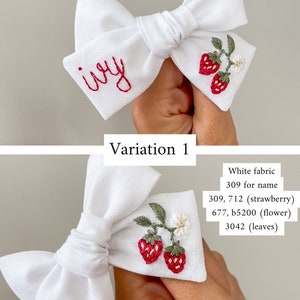 Op de afbeelding: Witte stoffen strik met rode geborduurde tekst "ivy" en een aardbeienontwerp. De strik heeft twee rode aardbeien, groene bladeren en een kleine witte bloem. Een charmant haaraccessoire.