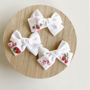 Op de afbeelding: Drie witte stoffen haarstrikken met geborduurde aardbeienontwerpen en namen. E&eacute;n strik heeft "Ada" geborduurd met een aardbei, een andere "Ivy" en de derde "Mary" met drie aardbeien. De strikken zijn gerangschikt op een licht houten oppervlak.