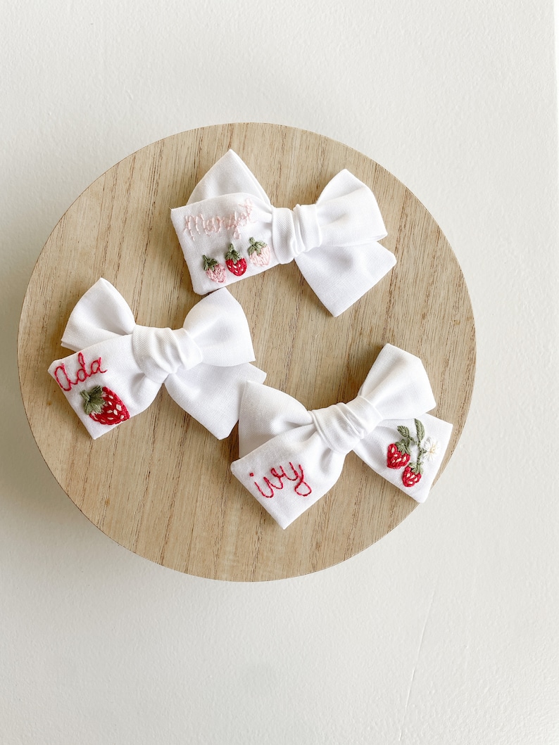 Op de afbeelding: Drie witte stoffen haarstrikken met geborduurde aardbeienontwerpen en namen. E&eacute;n strik heeft "Ada" geborduurd met een aardbei, een andere "Ivy" en de derde "Mary" met drie aardbeien. De strikken zijn gerangschikt op een licht houten oppervlak.