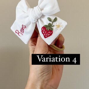 Op de afbeelding: Witte baby haarstrik met rood geborduurde naam en een aardbei ontwerp. De strik heeft een rood geborduurde aardbei, groene bladeren en een kleine witte bloem. De tekst "Variation 4" wordt weergegeven in een zwarte rechthoek.