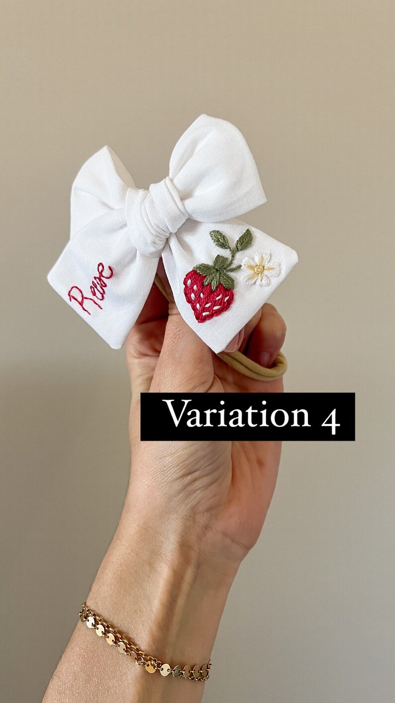 Op de afbeelding: Witte baby haarstrik met rood geborduurde naam en een aardbei ontwerp. De strik heeft een rood geborduurde aardbei, groene bladeren en een kleine witte bloem. De tekst "Variation 4" wordt weergegeven in een zwarte rechthoek.