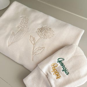May include: A cream-colored baby hat and a matching swaddle blanket with embroidered flowers and the name "Georgia Poppy" in green and yellow thread.