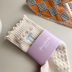 Op de afbeelding: Crèmekleurige sokken met het woord "mama" geborduurd in goudkleurig garen en een blauwe strik. De sokken hebben een geribbeld ontwerp en een lavendelkleurige band met de tekst "jus love MORE". Een baby-slab met een bruin en zwart ruitpatroon is ook zichtbaar.
