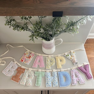 May include: A fabric banner spelling out HAPPY BIRTHDAY with individual stuffed letters in various patterns and colors, including floral, gingham, and striped prints. Each letter is attached to a cream-colored ribbon, creating a two-row display.