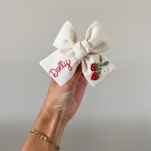 Op de afbeelding: Een cr&egrave;mekleurige stoffen strik met de naam "Dolly" in rode letters geborduurd. De strik is versierd met geborduurde aardbeien en bloemen. Dit handgemaakte haaraccessoire is ideaal om een vleugje charme aan elk kapsel toe te voegen.