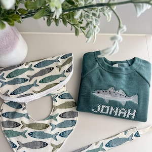 May include: A baby gift set featuring a green sweatshirt embroidered with a fish and the name "JONAH", two bibs, and a pacifier clip. The bibs and clip have a repeating fish pattern in shades of blue and green on a white background.