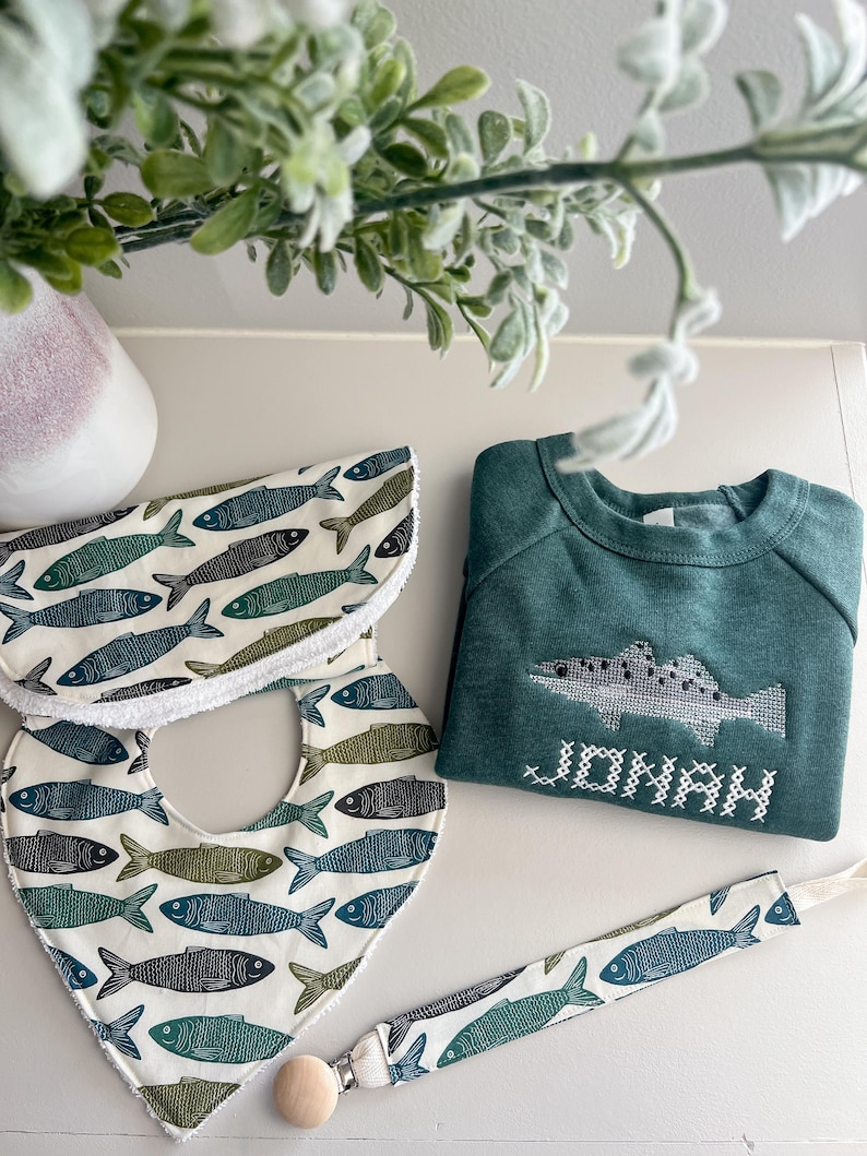 May include: A baby gift set featuring a green sweatshirt embroidered with a fish and the name "JONAH", two bibs, and a pacifier clip. The bibs and clip have a repeating fish pattern in shades of blue and green on a white background.