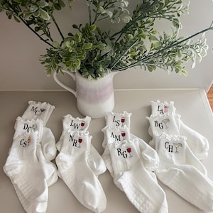 Könnte beinhalten: Weiße Knöchelsocken mit Rüschen, jede mit Initialen und einem Cocktail- oder Weinglas-Design bestickt. Eine Vase mit Grünzeug befindet sich im Hintergrund. Die Socken sind auf einer weißen Oberfläche angeordnet.