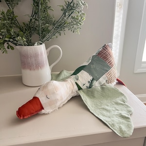 May include: A plush goose toy with a patchwork body, light green wings, and an orange beak. The goose has a cream-colored head with a gingham pattern. A white and pink pitcher with greenery is in the background.