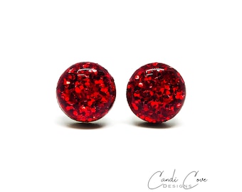 Può includere: Un paio di orecchini a bottone con glitter rosso. Gli orecchini sono rotondi e hanno una finitura lucida e scintillante.
