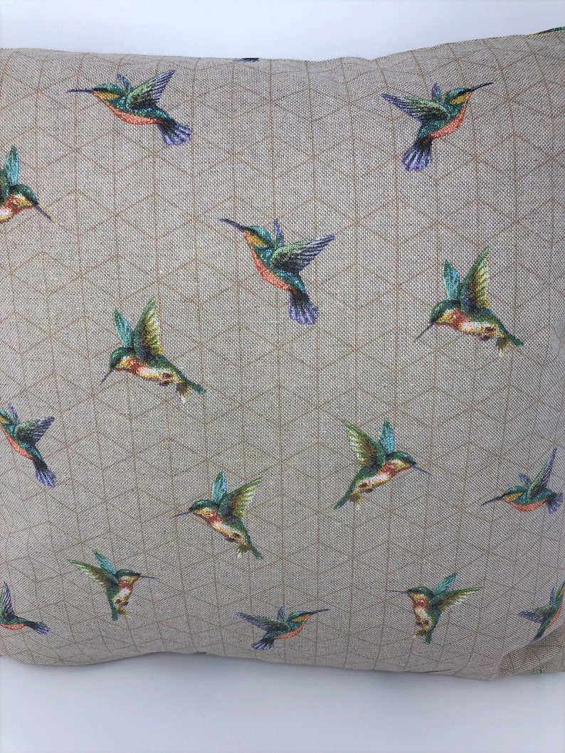 May include: A square cushion featuring a repeating pattern of hummingbirds in flight. The birds are green, orange, and purple. The background is a light tan fabric with a geometric design.