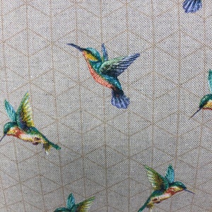 May include: A close-up of a fabric featuring a repeating pattern of hummingbirds. The birds are shown in flight with green, blue, orange, and yellow feathers. The background is a light beige with a subtle grid design.