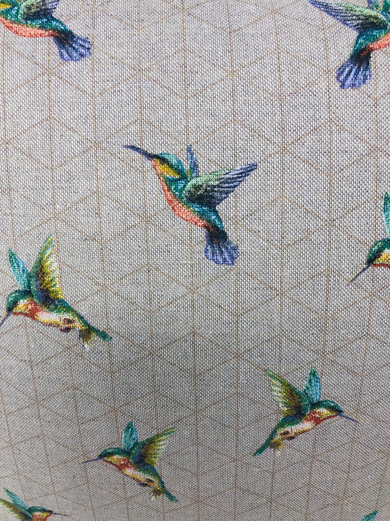 May include: A close-up of a fabric featuring a repeating pattern of hummingbirds. The birds are shown in flight with green, blue, orange, and yellow feathers. The background is a light beige with a subtle grid design.