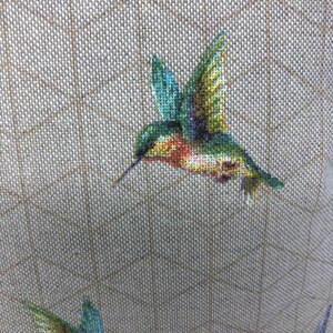May include: A close-up of a cross-stitch design showing multiple hummingbirds in flight. The birds are depicted with green, blue, yellow, and red hues. The background is a tan fabric with a grid pattern.