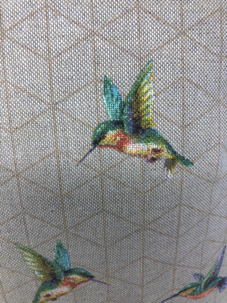 May include: A close-up of a cross-stitch design showing multiple hummingbirds in flight. The birds are depicted with green, blue, yellow, and red hues. The background is a tan fabric with a grid pattern.