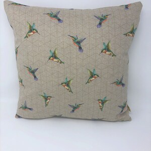 May include: Square cushion featuring a repeating pattern of colourful hummingbirds on a beige background. The birds have green, blue, orange, and red details. The cushion has a geometric design.