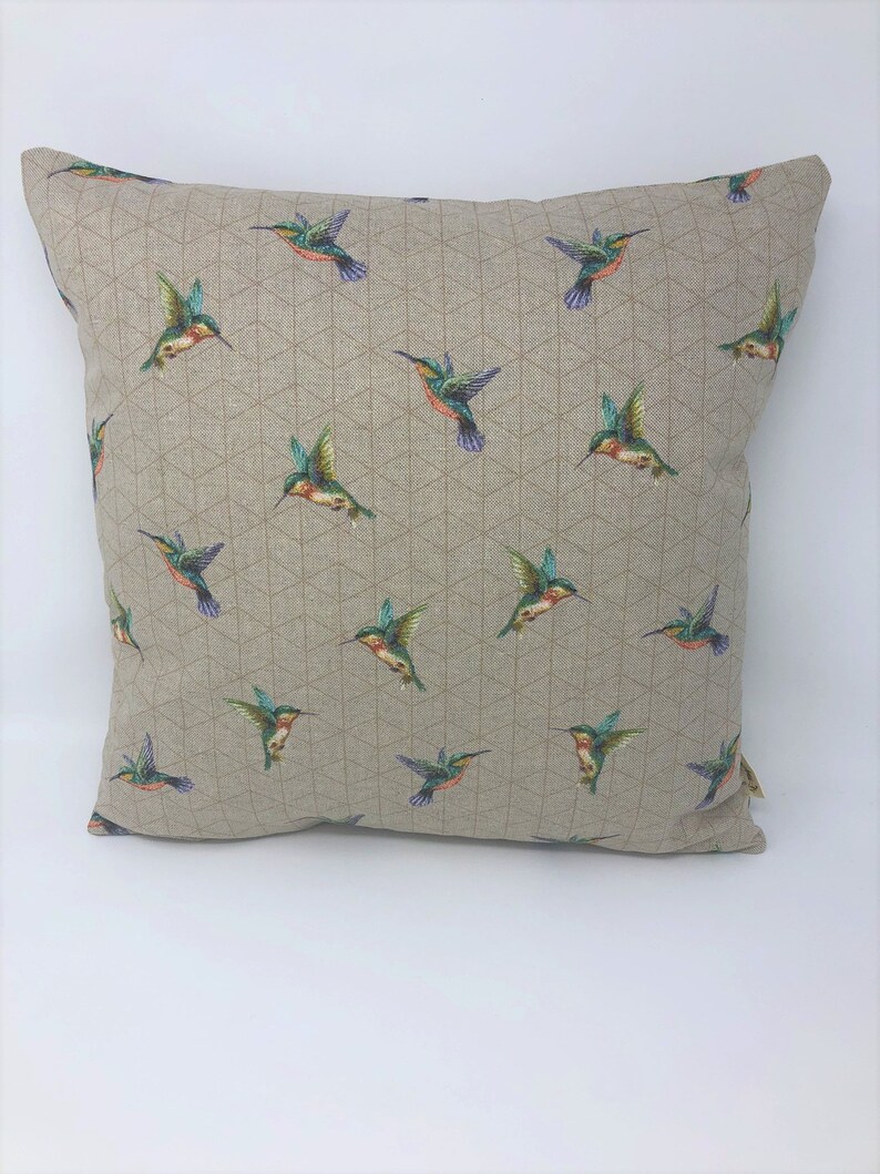May include: Square cushion featuring a repeating pattern of colourful hummingbirds on a beige background. The birds have green, blue, orange, and red details. The cushion has a geometric design.