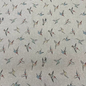 May include: Pale grey fabric with a repeating pattern of hummingbirds in flight. The birds are illustrated in shades of grey, blue, red, and green. The fabric has a soft, textured appearance, suitable for various sewing projects.