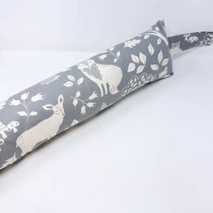 May include: A grey fabric doorstop with a woodland animal pattern. The design features white deer, foxes, and floral elements. A matching fabric handle is attached to the end of the doorstop.