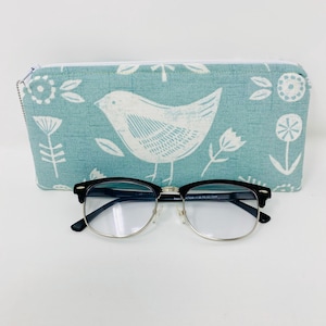 May include: A light blue fabric glasses case with a white bird and floral pattern. The case is closed and a pair of black framed glasses are in front of it.