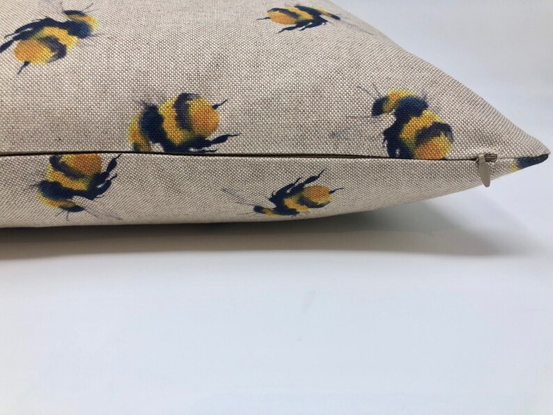 May include: A beige linen pillow with a zipper closure features a repeating pattern of yellow and black bumblebees.