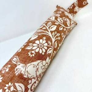 May include: A brown and white fabric doorstop with a floral and rabbit design. The cylindrical doorstop has a handle and is made of textured fabric. The pattern includes flowers, leaves, and a rabbit silhouette.