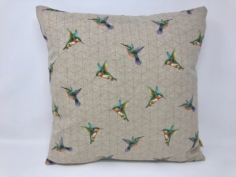 May include: Square cushion featuring a repeating pattern of hummingbirds. The birds are depicted in green, blue, and red against a neutral background with a geometric design. The cushion is square in shape.