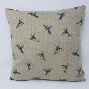 May include: Square cushion featuring a repeating pattern of hummingbirds. The birds are depicted with green, blue, and orange feathers, set against a neutral background with a geometric design. The cushion is square.