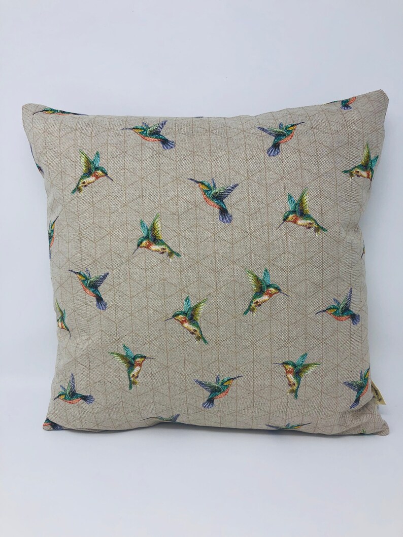 May include: Square cushion featuring a repeating pattern of hummingbirds. The birds are depicted with green, blue, and orange feathers, set against a neutral background with a geometric design. The cushion is square.