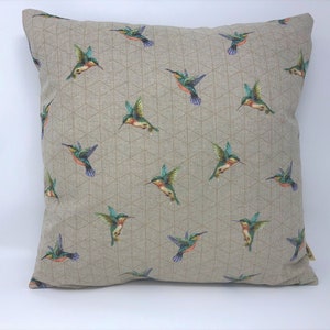 May include: Square cushion featuring a repeating pattern of hummingbirds. The birds are depicted in flight with green, blue, orange, and purple plumage. The cushion has a neutral background with a geometric design.