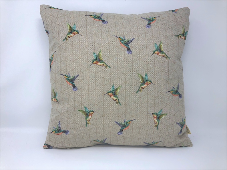 May include: Square cushion featuring a repeating pattern of hummingbirds. The birds are depicted in flight with green, blue, orange, and purple plumage. The cushion has a neutral background with a geometric design.