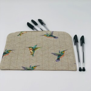 May include: A beige pencil case featuring a geometric design and vibrant hummingbird illustrations. Several black pens are visible, some inside the case and others beside it. The case has a zip closure and a rectangular shape.