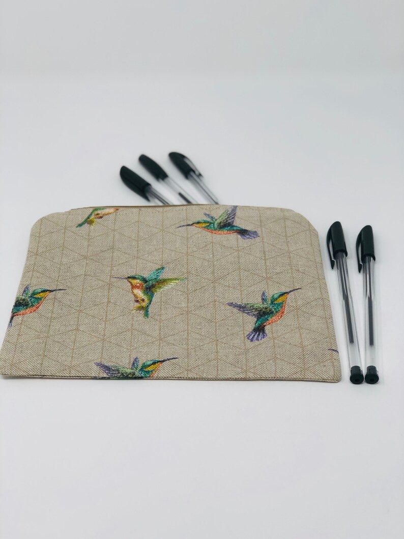 May include: A beige pencil case featuring a geometric design and vibrant hummingbird illustrations. Several black pens are visible, some inside the case and others beside it. The case has a zip closure and a rectangular shape.