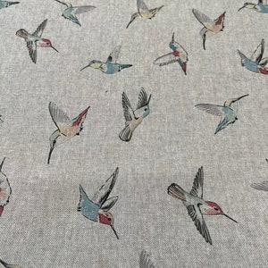 May include: A light grey fabric with a repeating pattern of hummingbirds in flight. The birds feature a variety of colours, including red, blue, green, and yellow. The fabric has a textured appearance.