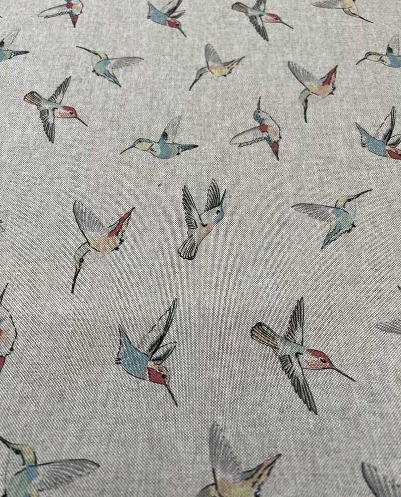 May include: A light grey fabric with a repeating pattern of hummingbirds in flight. The birds feature a variety of colours, including red, blue, green, and yellow. The fabric has a textured appearance.