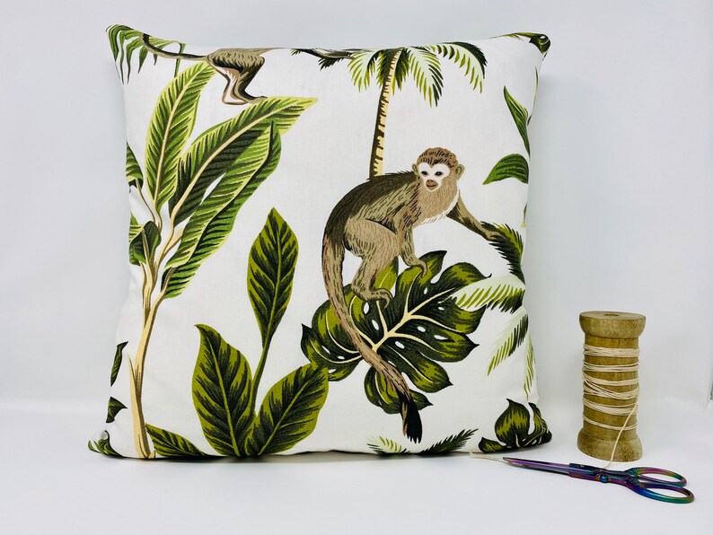 May include: White throw pillow with a tropical print featuring monkeys and palm leaves. The pillow has a green, brown, and white color scheme.