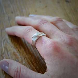 May include: A silver ring with a geometric design. The ring is worn on the middle finger of a hand.
