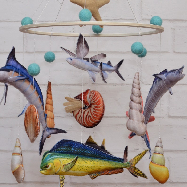 May include: A colorful mobile with a variety of sea creatures, including a marlin, a sailfish, a dolphin, a nautilus, and seashells. The mobile is made of felt and fabric and is suspended from a wooden ring.