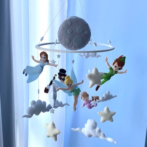 Peut inclure: Un mobile bébé en feutre blanc avec une lune grise, des nuages, des étoiles et des personnages en feutre, dont Peter Pan, la fée Clochette et une fée.