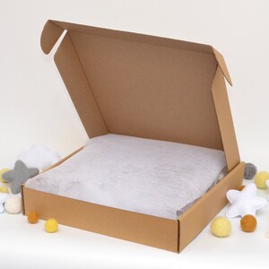 May include: A brown cardboard box with the top open, revealing white tissue paper inside. The box is sitting on a white surface with yellow and gray pom-poms scattered around it.
