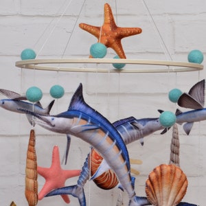May include: A wooden mobile with a variety of sea-themed felt toys, including a marlin, a flying fish, a starfish, a seashell, and a coral. The mobile is suspended from a wooden ring and has turquoise felt balls hanging from it.