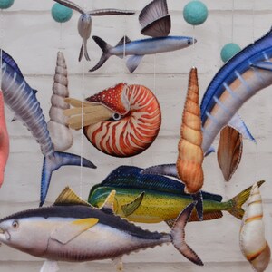 May include: A colorful mobile featuring a variety of sea creatures, including a nautilus, a mahi-mahi, a tuna, a flying fish, and a marlin. The mobile is made of felt and is suspended from a white ceiling.