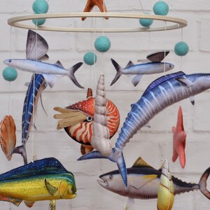 May include: A colorful mobile with a variety of sea creatures, including a marlin, a dolphin, a tuna, a nautilus, and a flying fish. The mobile is made of felt and is suspended from a wooden ring.