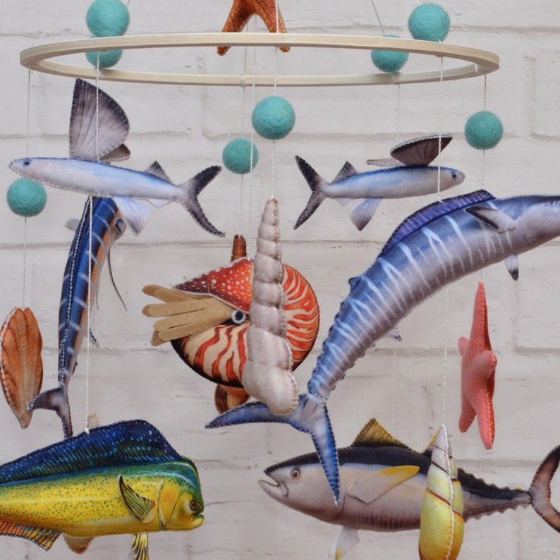 May include: A colorful mobile with a variety of sea creatures, including a marlin, a dolphin, a tuna, a nautilus, and a flying fish. The mobile is made of felt and is suspended from a wooden ring.