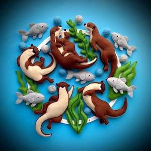 May include: A felt mobile with brown otters and gray fish hanging from a white ring. The otters are surrounded by green felt seaweed and white felt balls.