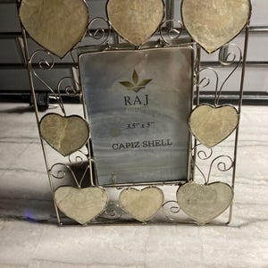 Puede incluir: Un marco de fotos rectangular de metal plateado con seis detalles en forma de corazón hechos de concha capiz iridiscente. El inserto central muestra el texto RAJ Collection, 3.5" x 5" y CAPIZ SHELL. El marco tiene adornos de alambre en espiral.