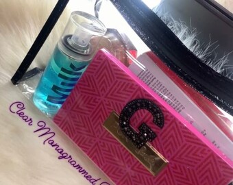 May include: Clear travel bag with a black zipper and a pink monogrammed pouch with a large rhinestone G inside. The bag is filled with various items, including a blue spray bottle with the word "Milk" on it.