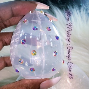 Puede incluir: Una caja de esponja de maquillaje de plástico transparente con un diseño decorativo de diamantes de imitación iridiscentes. La caja está abierta y la mitad superior está siendo sostenida por una mano.