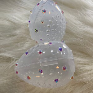 May include: Two clear plastic egg-shaped containers with a textured surface and iridescent rhinestones. The containers have a small opening on the side.