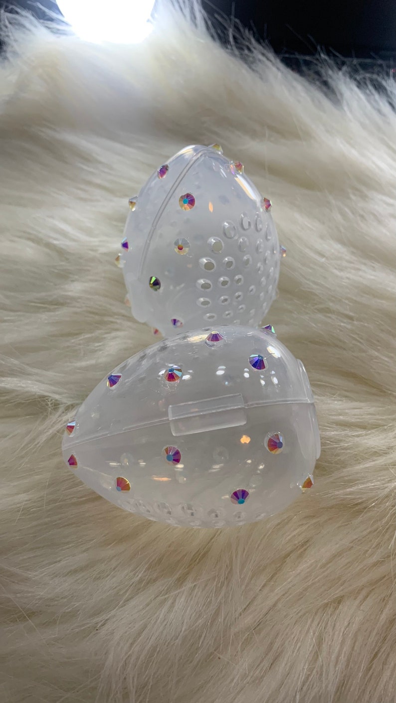 May include: Two clear plastic egg-shaped containers with a textured surface and iridescent rhinestones. The containers have a small opening on the side.